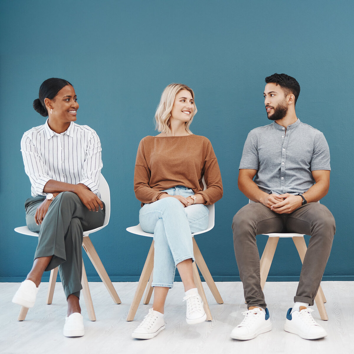 Three people sit on chairs in front of a bright blue wall. They are looking at each other.